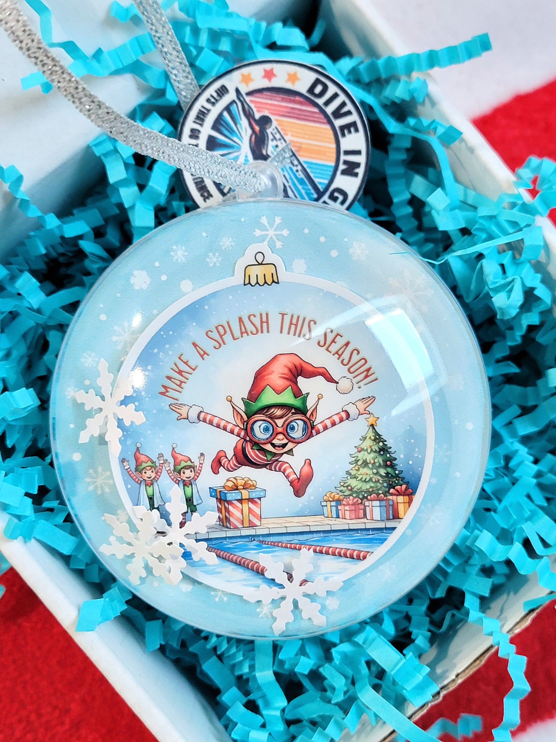 Decorative ornament with elf design and text in a gift box with blue crinkle paper.
